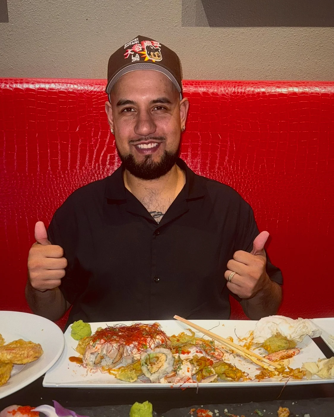 Brandon of Loco Eatz giving two thumbs up from a red booth, a sushi spread on the table